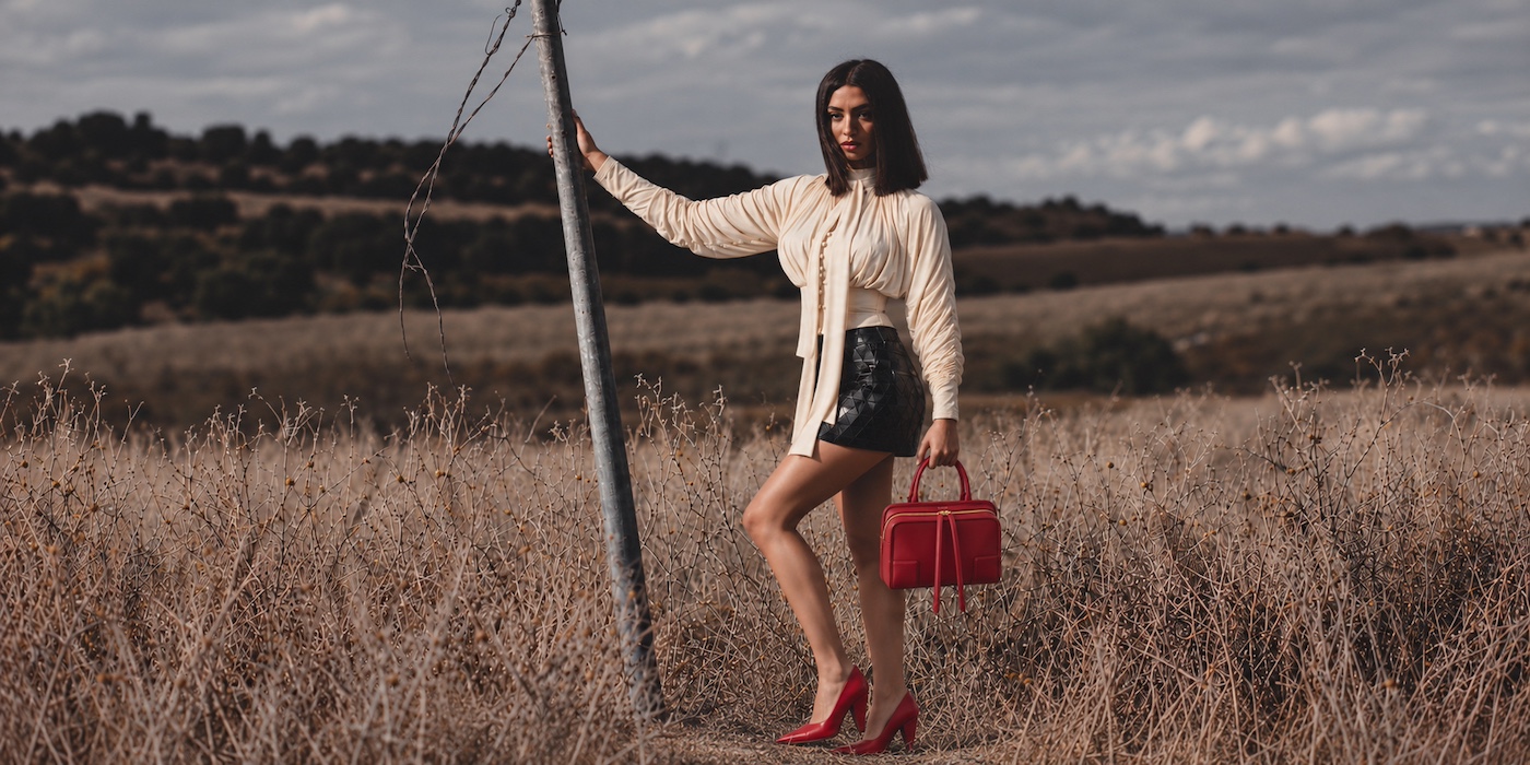 Model in a cream blouse and black skirt holding a red handbag in a dry field