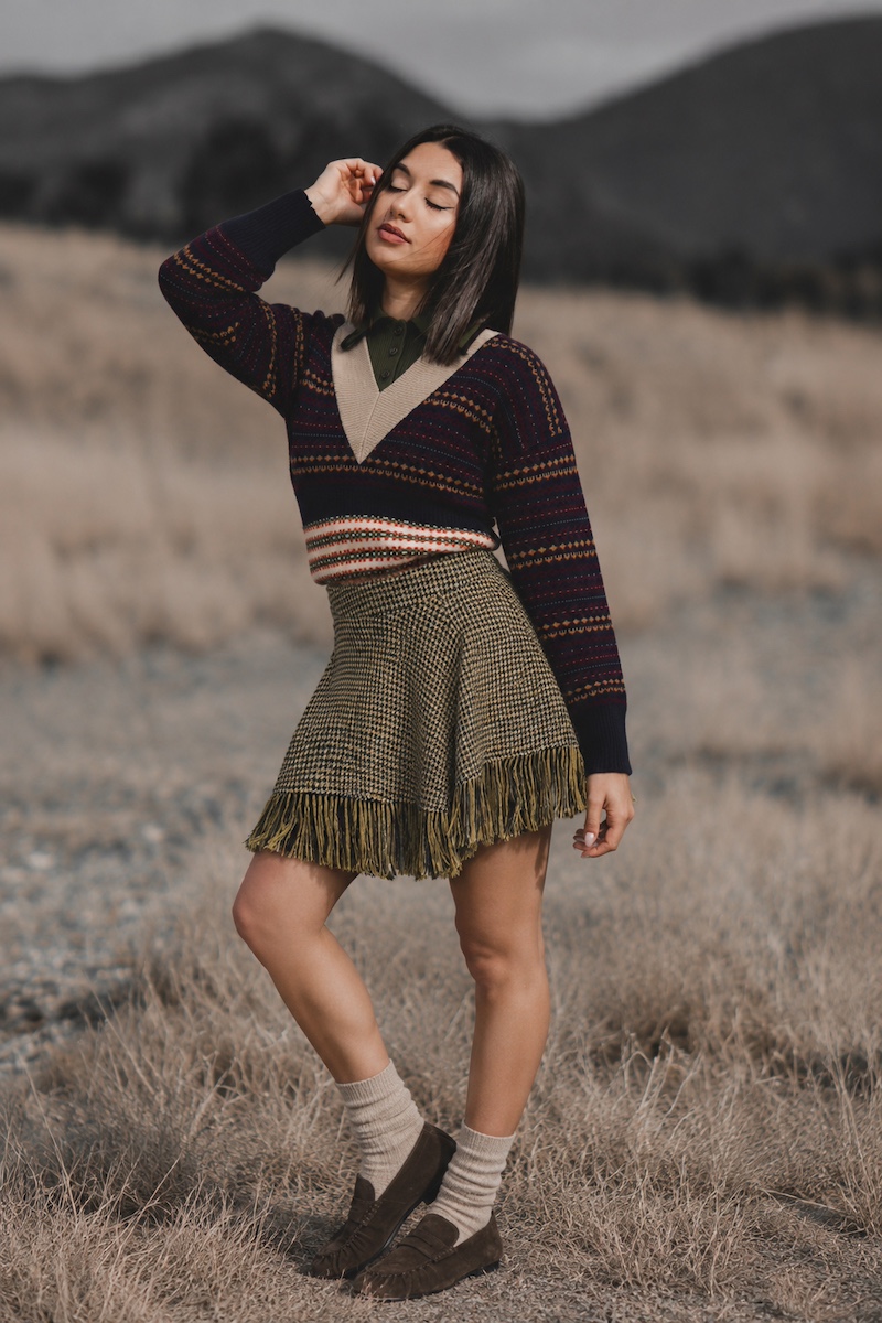 Model in a fair isle knit, fringed houndstooth skirt and brown loafers in a dry field